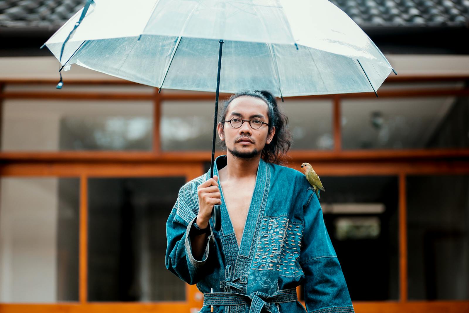 Man in denim kimono holding an umbrella with a bird perched, street style portrait.